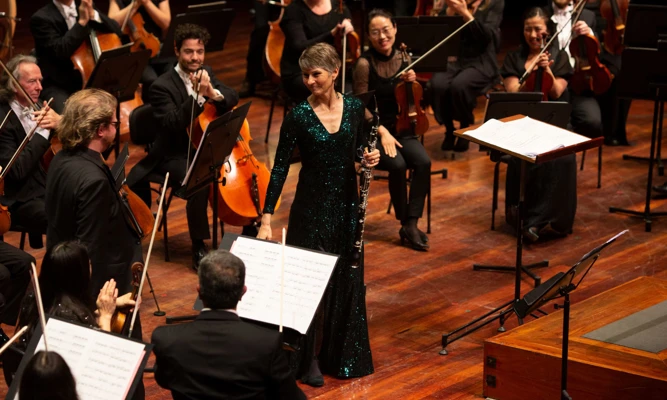 Soloist Leanne Glover acknowledges applause while standing among orchestra musicians on stage, wearing a sparkly green gown.