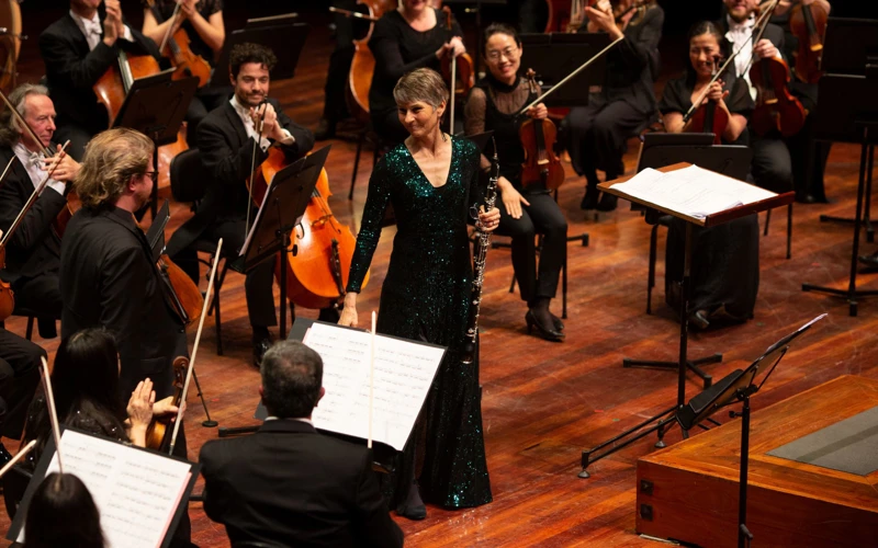 Soloist Leanne Glover acknowledges applause while standing among orchestra musicians on stage, wearing a sparkly green gown.