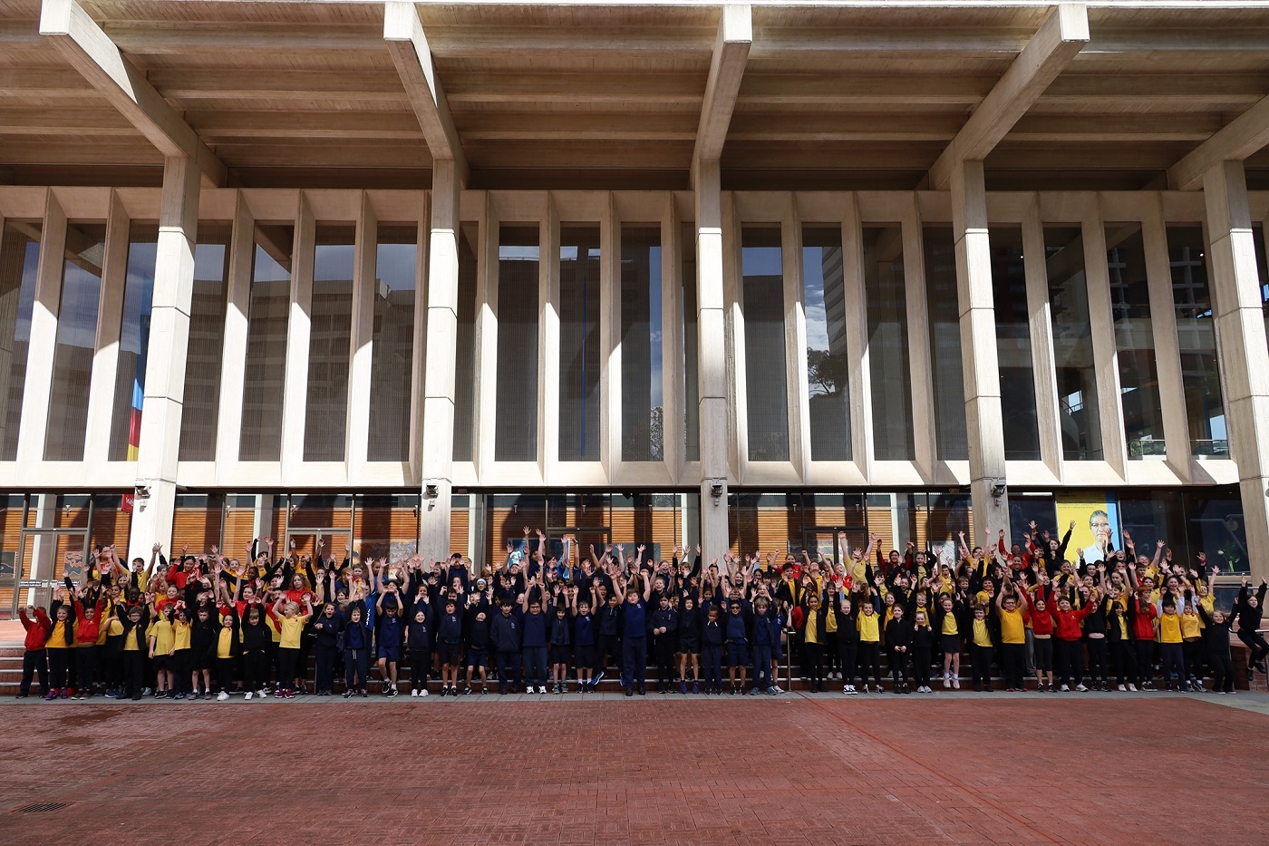 A large group of primary school students smiling with their hands raised in the air in front of Perth Concert Hall.