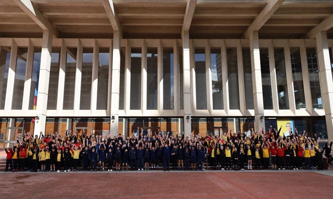 A large group of primary school students smiling with their hands raised in the air in front of Perth Concert Hall.