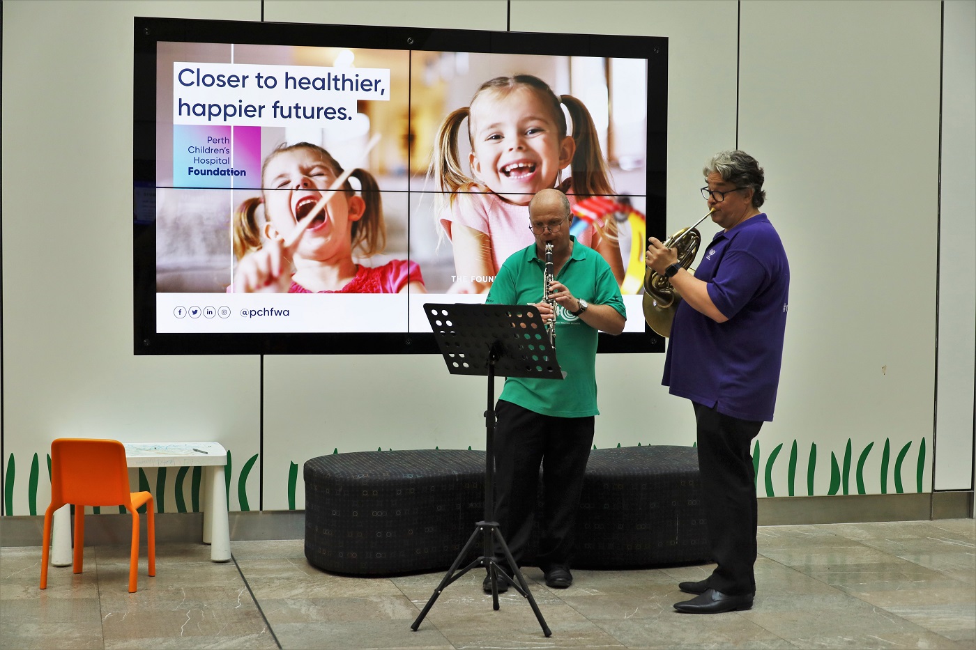 A clarinettist and horn player perform together at a children's hospital, reading from sheet music on stands.