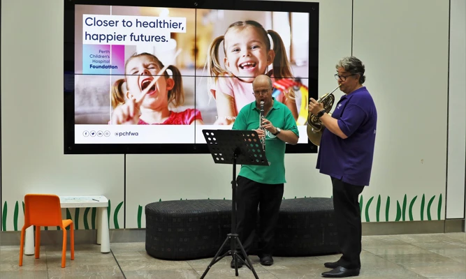 A clarinettist and horn player perform together at a children's hospital, reading from sheet music on stands.