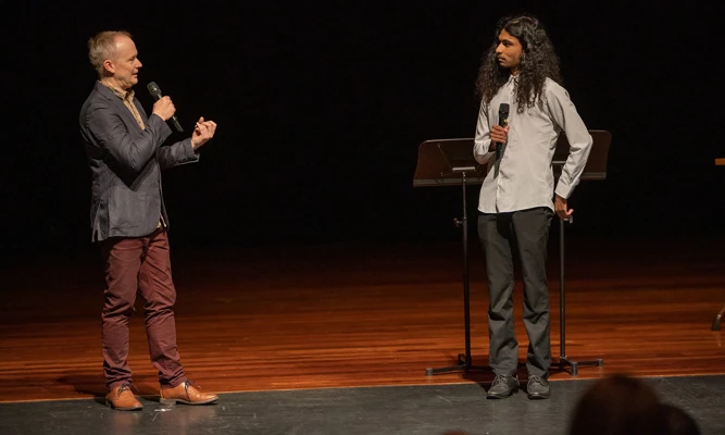 James Ledger and Victor Arul stand on stage for live discussion, speaking into microphones before an audience.