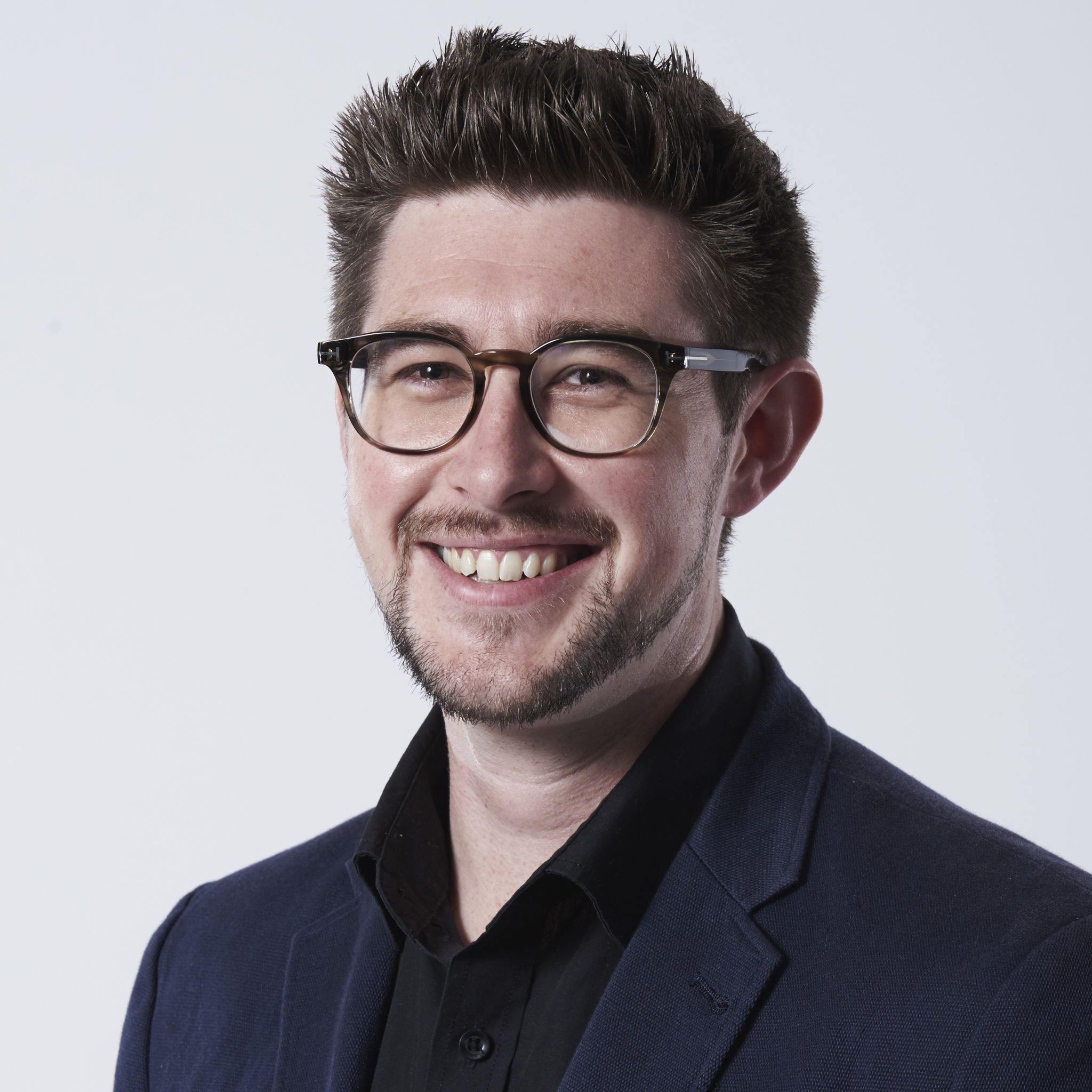 Headshot of Hugh Lydon wearing a navy suit jacket, black shirt and glasses.