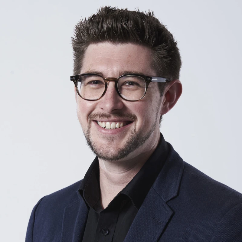 Headshot of Hugh Lydon wearing a navy suit jacket, black shirt and glasses.