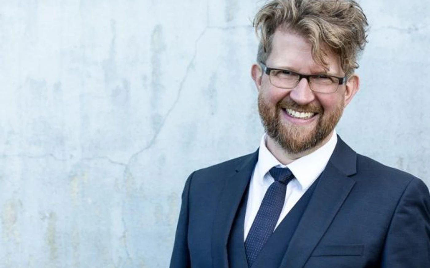 Headshot of Iain Grandage wearing a navy three-piece suit and tie with glasses.