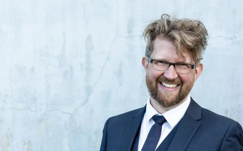 Headshot of Iain Grandage wearing a navy three-piece suit and tie with glasses.