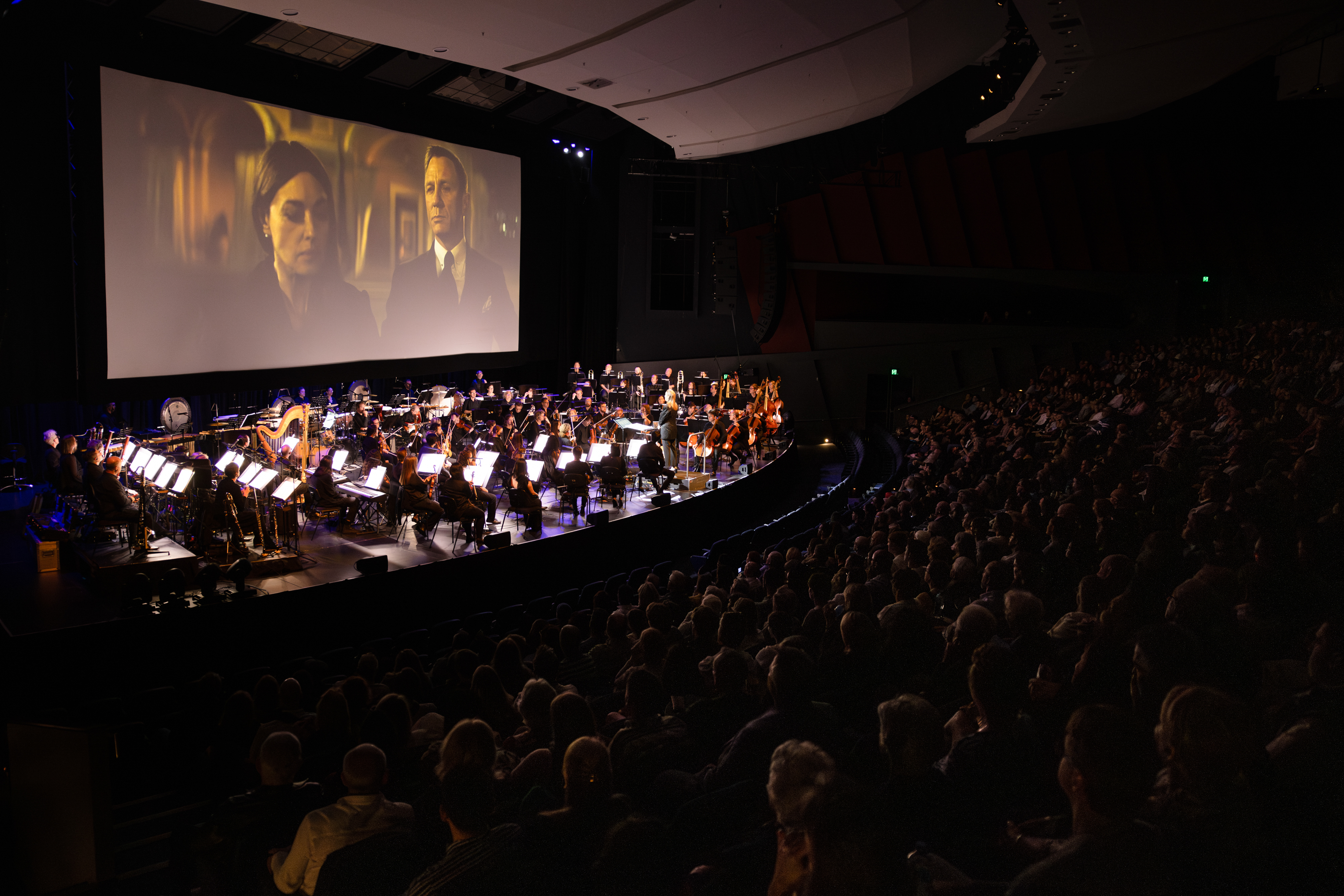 WASO performs on stage at the River Side Theatre, PCEC, with a large screen behind the orchestra displaying a James Bond film