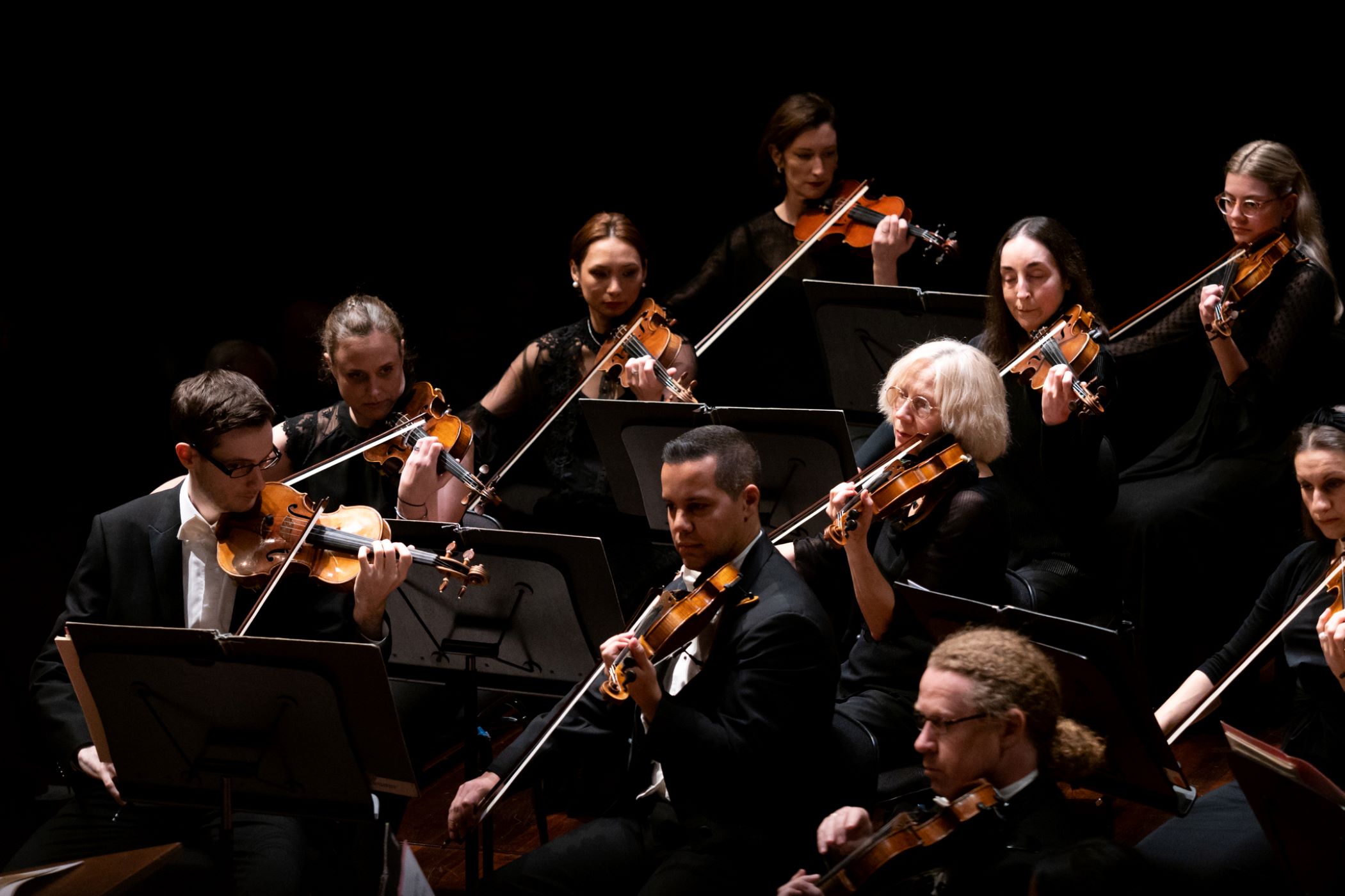 Violinists performing on stage in black formal wear, led by their concertmaster.