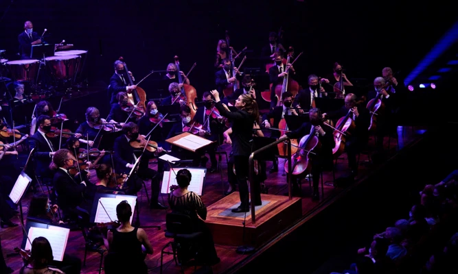 Jen Winley conducting an orchestra under dim purple stage lights.