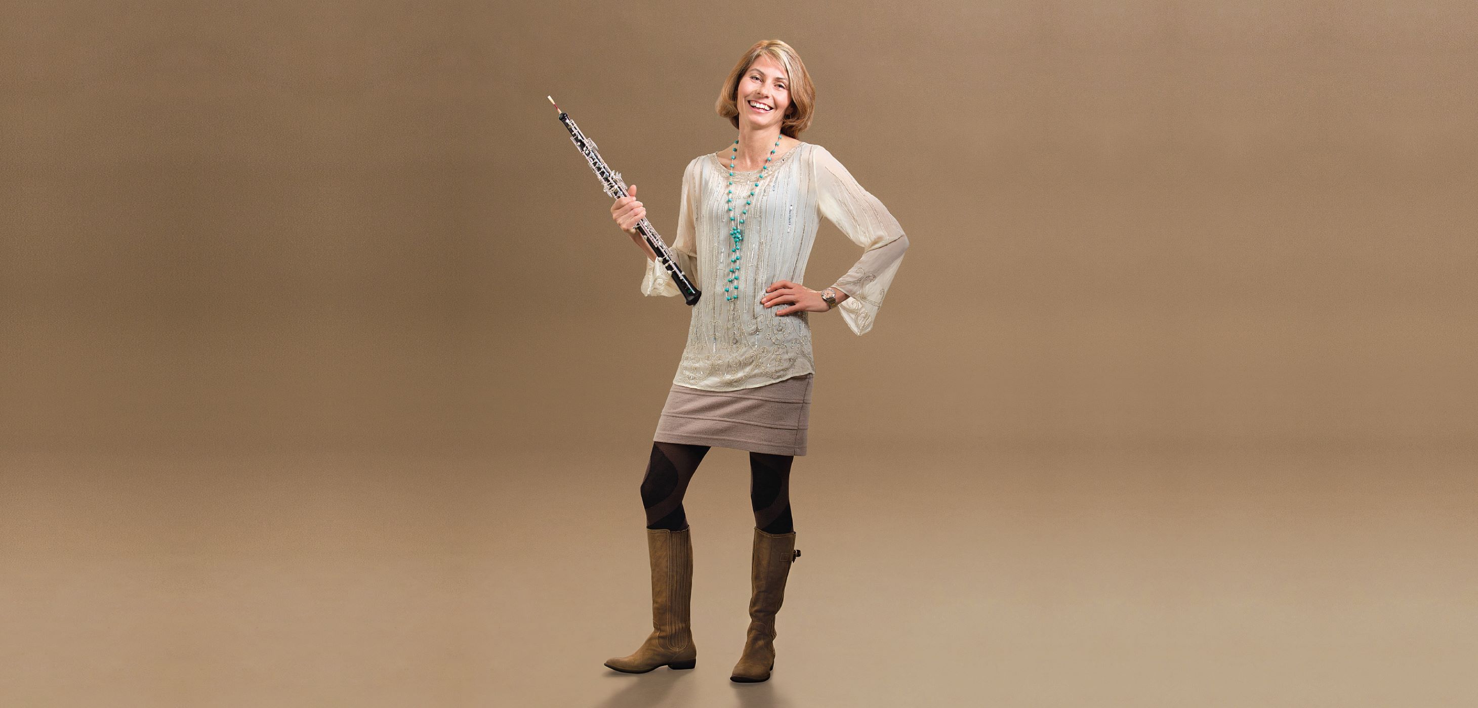 Full-length studio portrait of Leanne Glover wearing a long-sleeve dress and boots, holding an oboe.