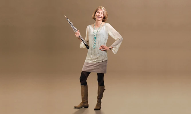 Full-length studio portrait of Leanne Glover wearing a long-sleeve dress and boots, holding an oboe.