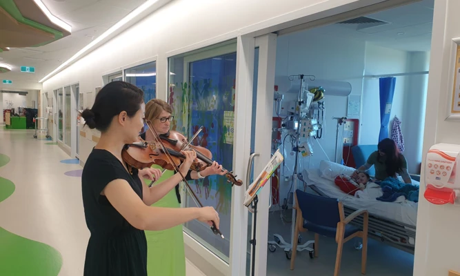 A violinist and violist performing together in a hospital corridor, while a young patient and her caretaker watch from bed.