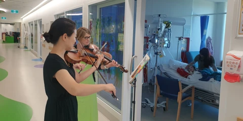 A violinist and violist performing together in a hospital corridor, while a young patient and her caretaker watch from bed.