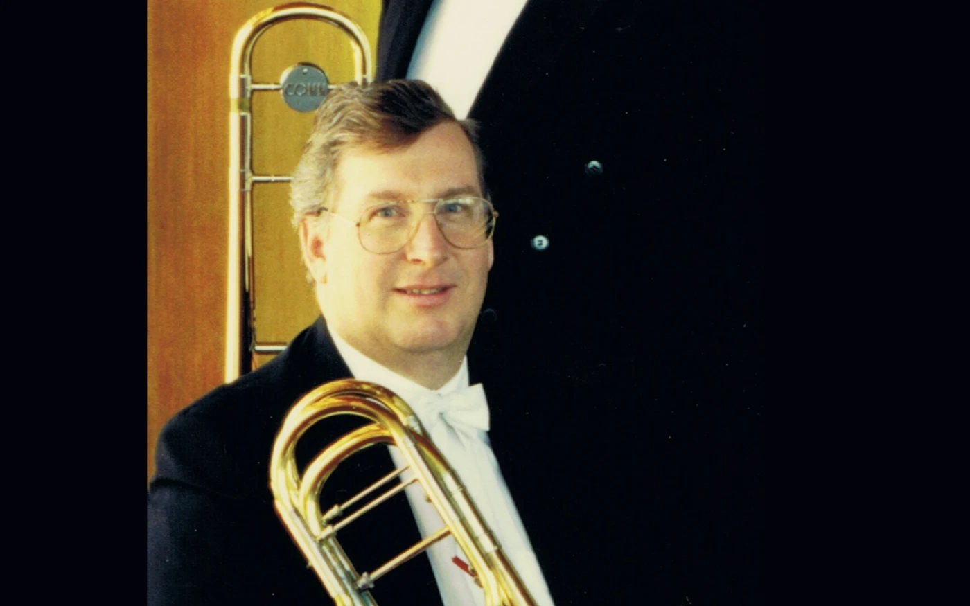 Headshot of Philip Holdsworth dressed in a formal suit, holding a trombone.