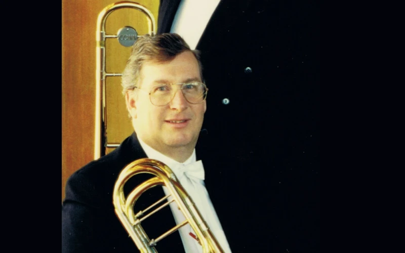 Headshot of Philip Holdsworth dressed in a formal suit, holding a trombone.
