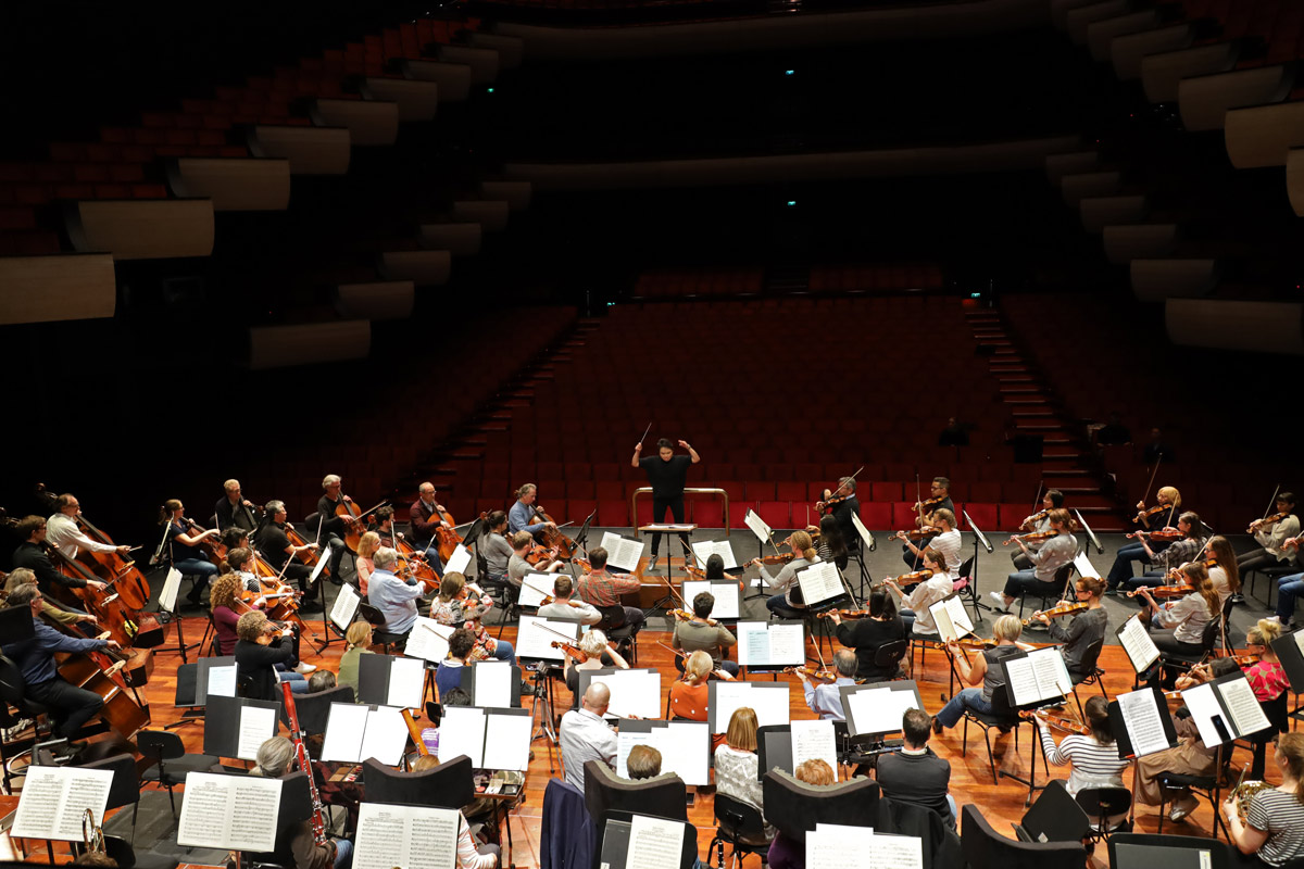 Elise Chong conducts an orchestra rehearsing on stage in an empty concert hall, dressed in casualwear.
