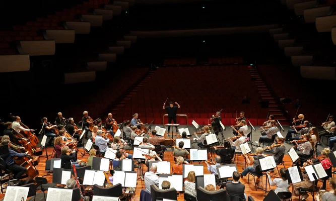 Elise Chong conducts an orchestra rehearsing on stage in an empty concert hall, dressed in casualwear.