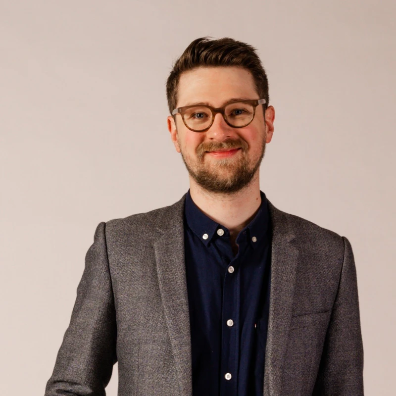Headshot of Dan Golding wearing a grey suit jacket, navy shirt and glasses.