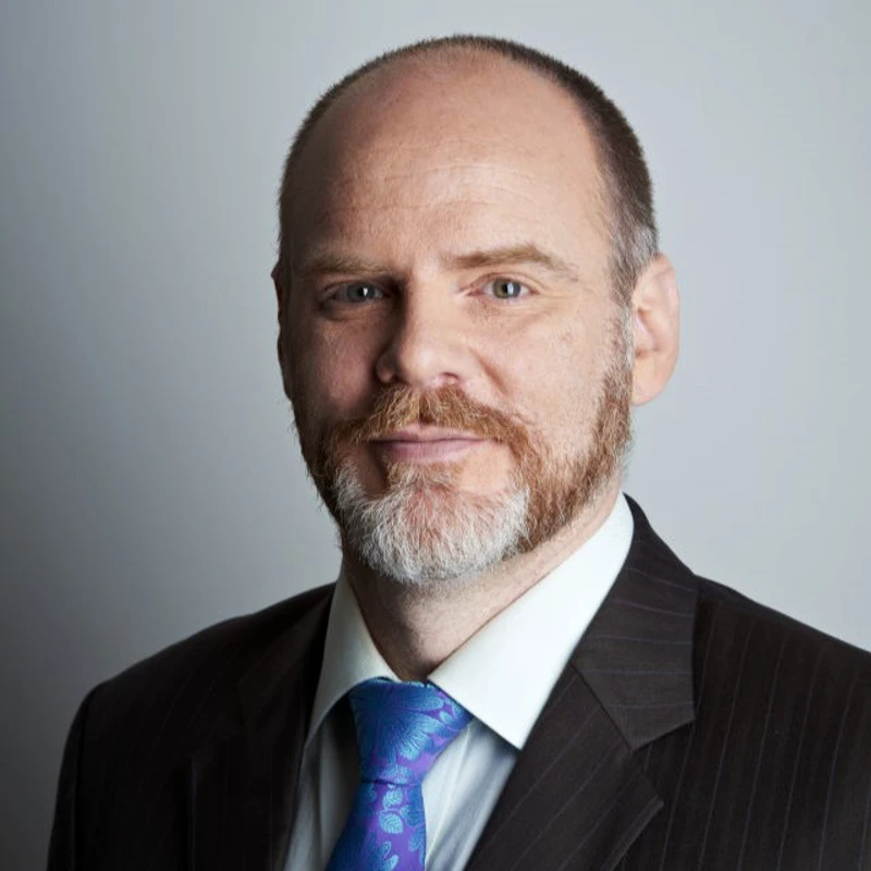 Headshot of James Clayton wearing a black pinstripe suit jacket, white shirt and blue floral tie.