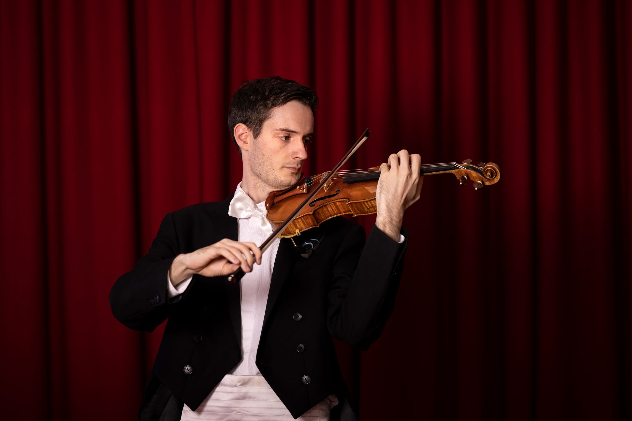 Riley Skevington playing a violin while dressed in a formal suit, in front of a red curtain backdrop.