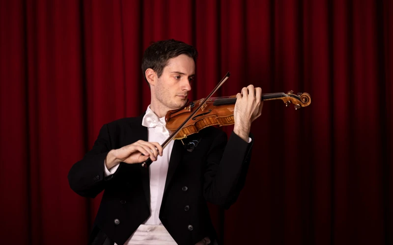 Riley Skevington playing a violin while dressed in a formal suit, in front of a red curtain backdrop.