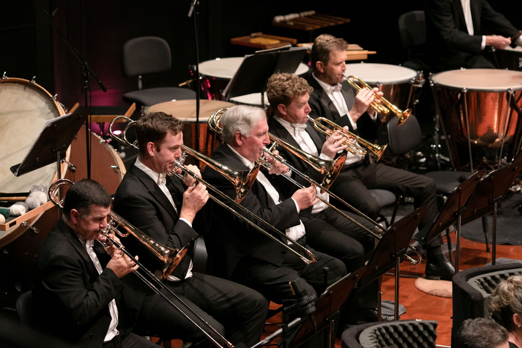Trumpet and trombone players performing on stage as part of an orchestra, dressed in formal concert attire.