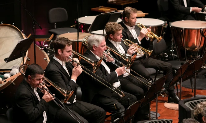 Trumpet and trombone players performing on stage as part of an orchestra, dressed in formal concert attire.