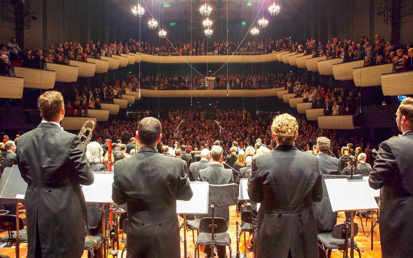 Orchestra members stand on a concert hall stage for applause, while a full audience gives a standing ovation.