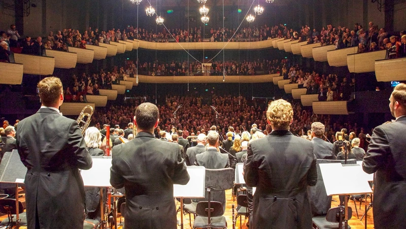 Orchestra members stand on a concert hall stage for applause, while a full audience gives a standing ovation.