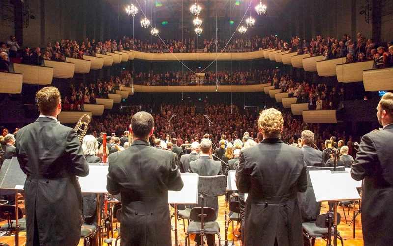 Orchestra members stand on a concert hall stage for applause, while a full audience gives a standing ovation.