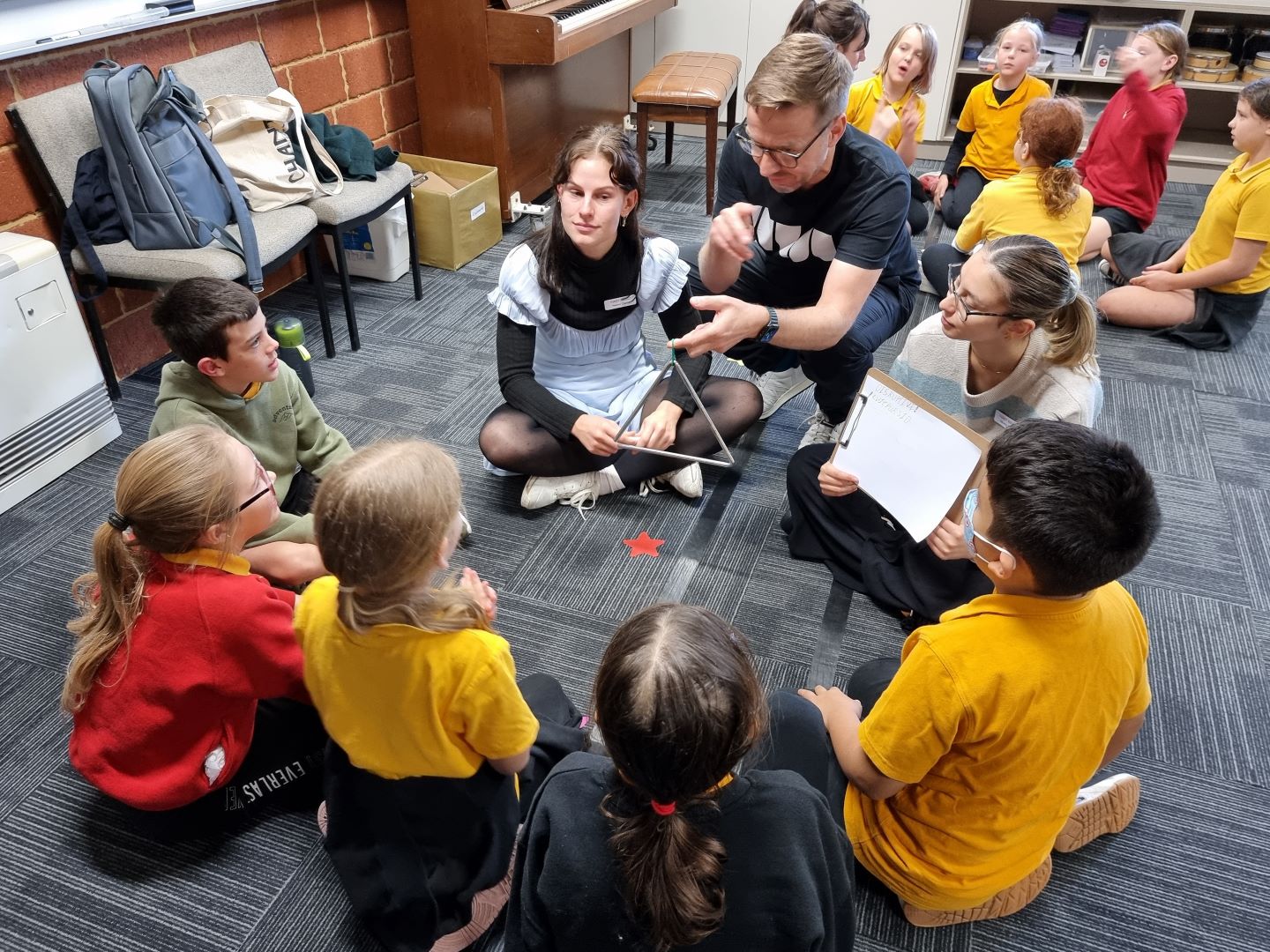 Students sit on the floor of a classroom watching a musician demonstrate playing the triangle