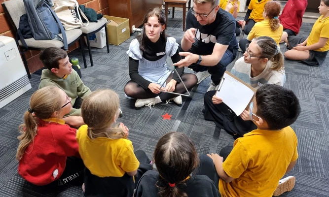 Students sit on the floor of a classroom watching a musician demonstrate playing the triangle