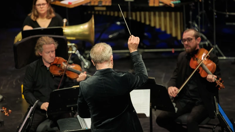 A conductor has his baton in the air as violinists and brass players play in front of him