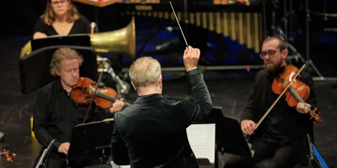 A conductor has his baton in the air as violinists and brass players play in front of him