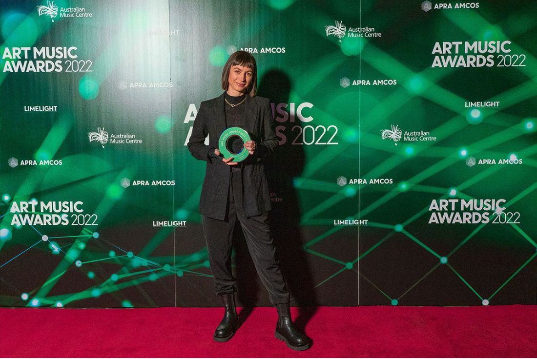 Olivia Davies poses with a trophy on the red carpet of the Art Music Awards 2022, wearing a black pantsuit and boots.