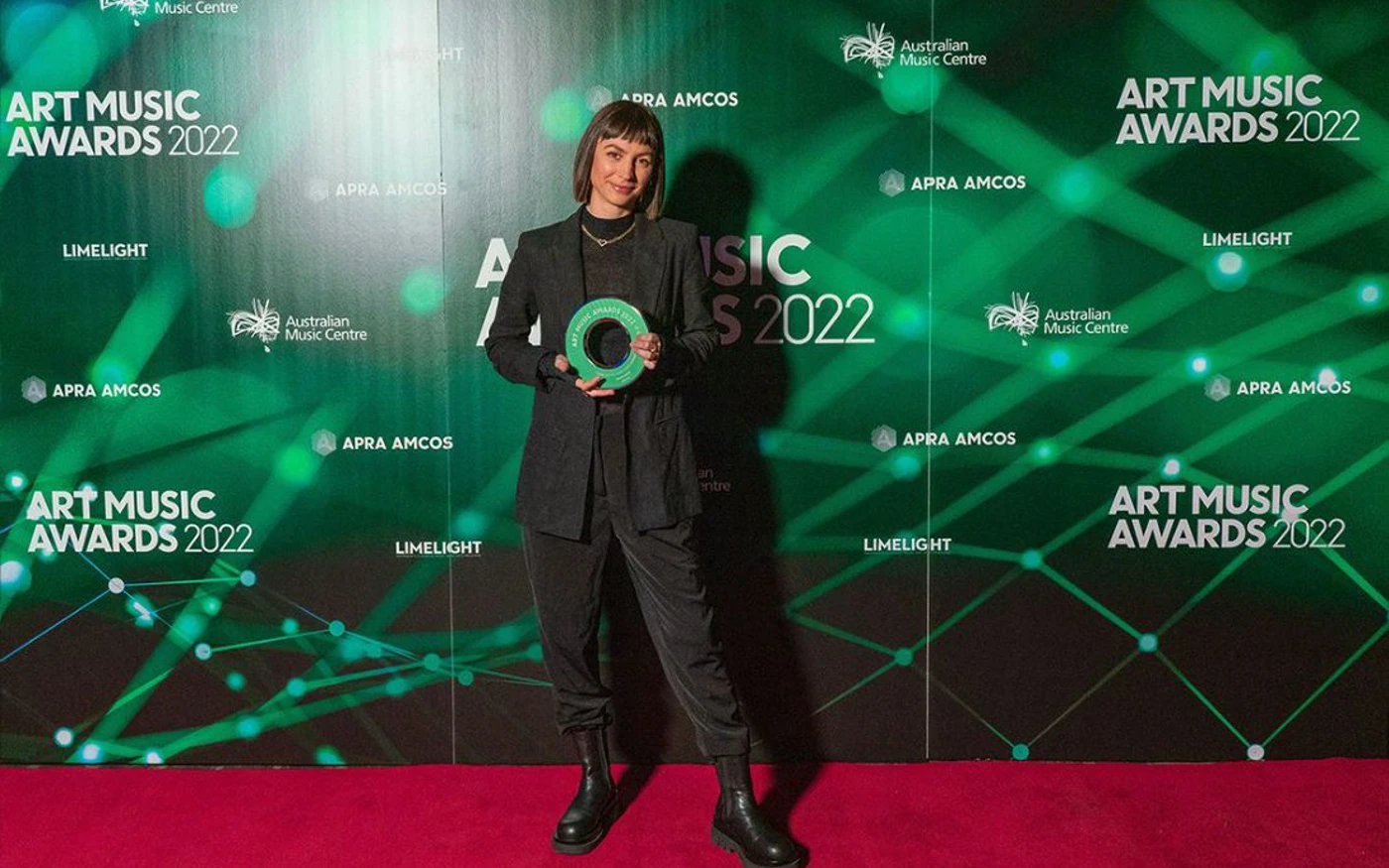 Olivia Davies poses with a trophy on the red carpet of the Art Music Awards 2022, wearing a black pantsuit and boots.