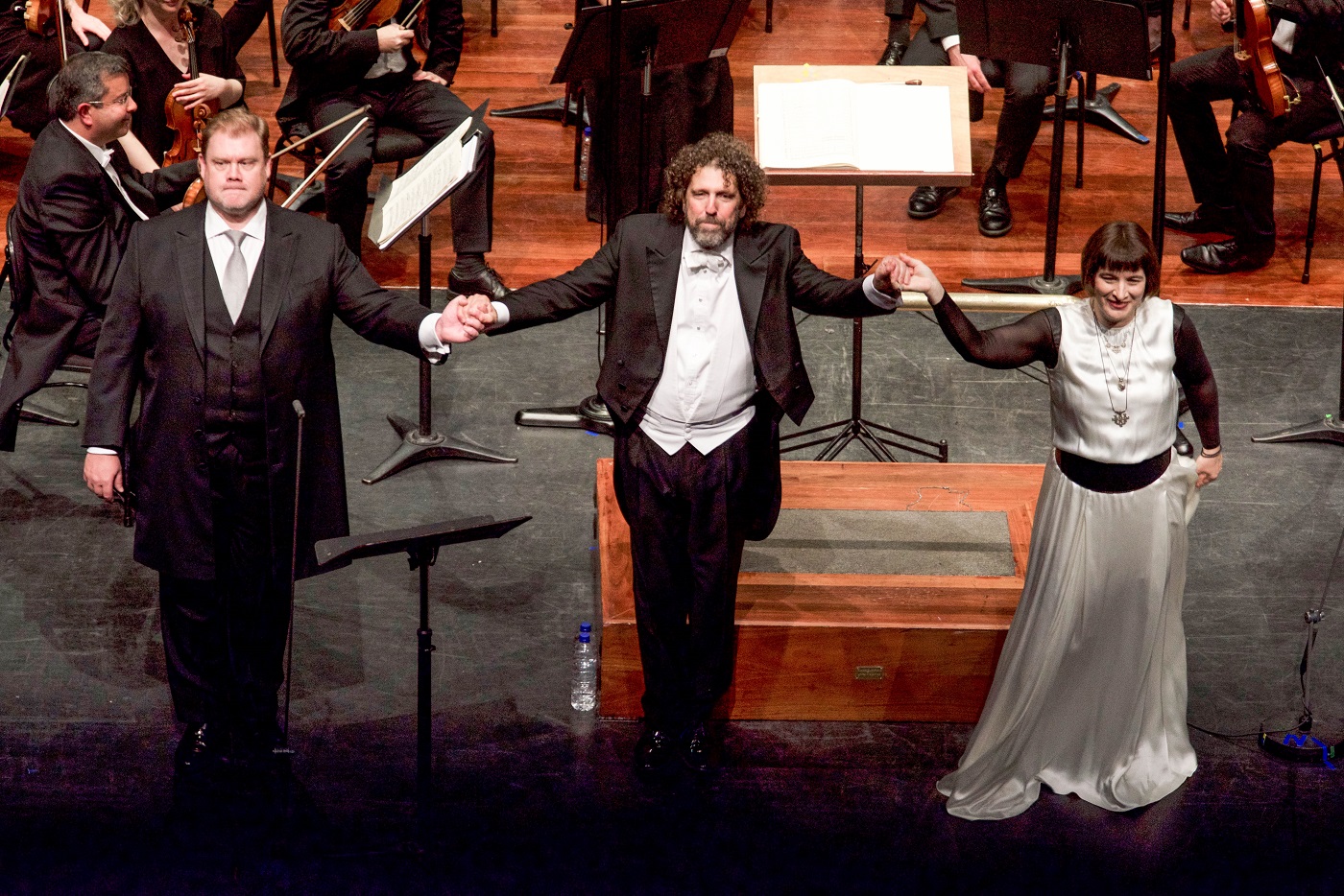 Two vocal soloists and a conductor stand hand-in-hand before taking a bow on stage.