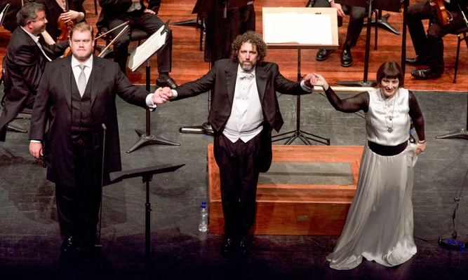 Two vocal soloists and a conductor stand hand-in-hand before taking a bow on stage.