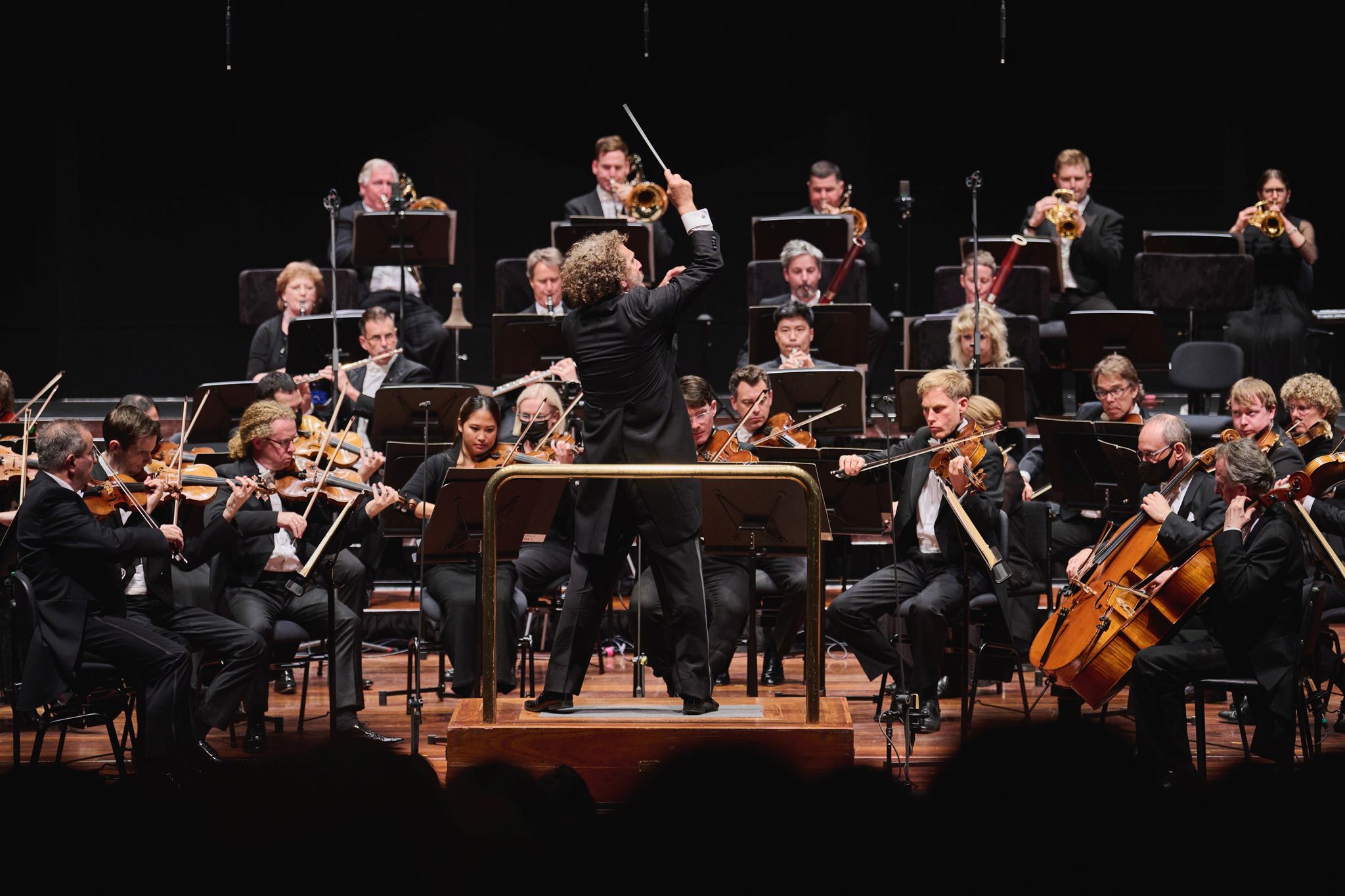 Asher Fisch conducts WASO with his back to the audience and one arm raised dramatically in the air. 