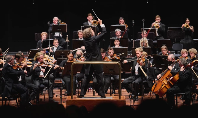 Asher Fisch conducts WASO with his back to the audience and one arm raised dramatically in the air.