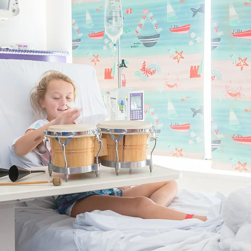 A young girl in a hospital bed plays a set of bongo drums