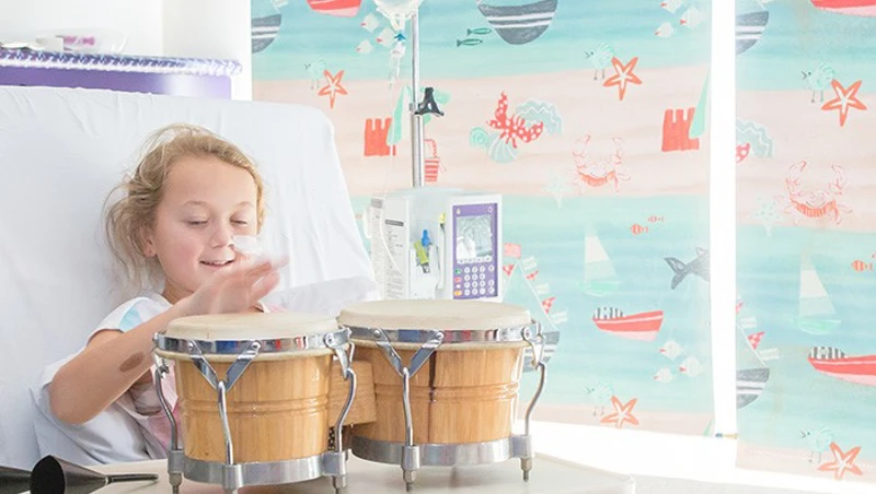 A young girl in a hospital bed plays a set of bongo drums