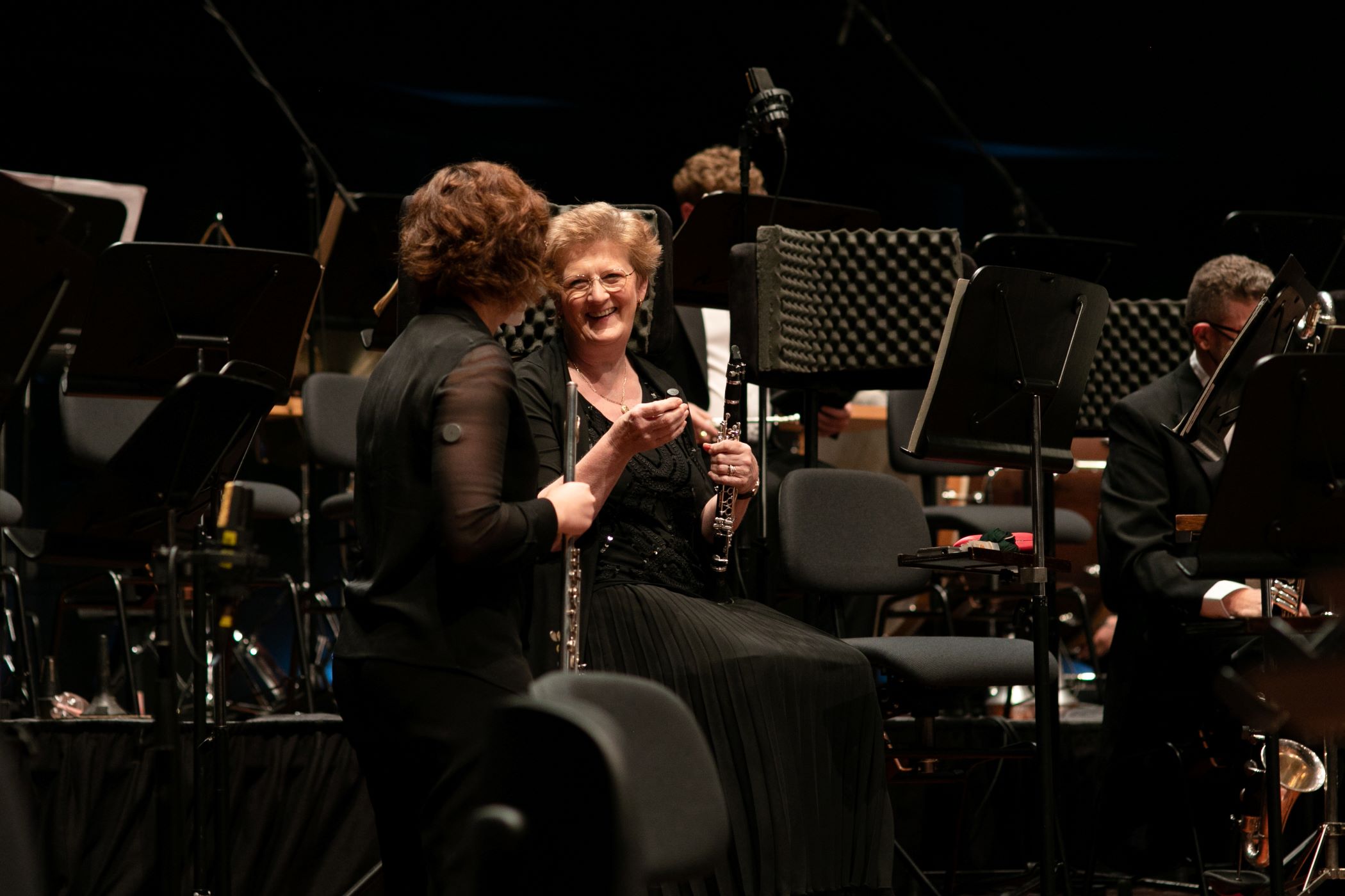 Two orchestra musicians holding woodwind instruments chat on stage among music stands and chairs.
