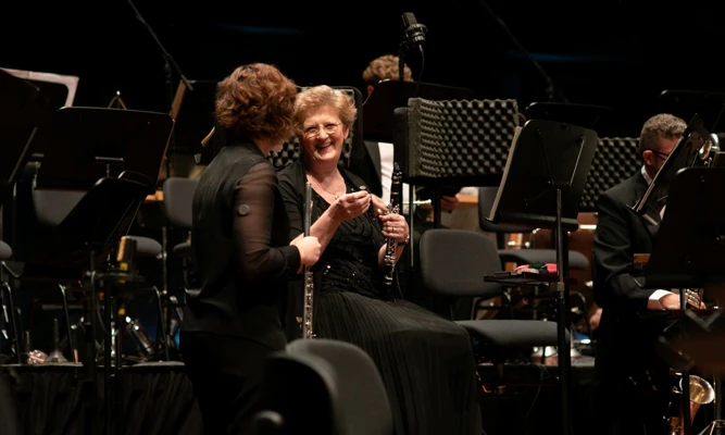 Two orchestra musicians holding woodwind instruments chat on stage among music stands and chairs.