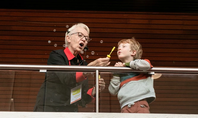 Theatre usher and young child stand on an indoor balcony, blowing soap bubbles together as bubbles float through the air.