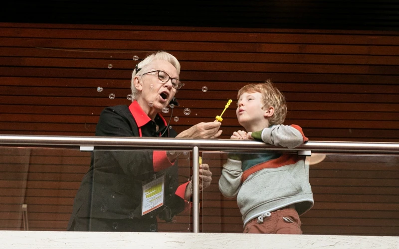 Theatre usher and young child stand on an indoor balcony, blowing soap bubbles together as bubbles float through the air.