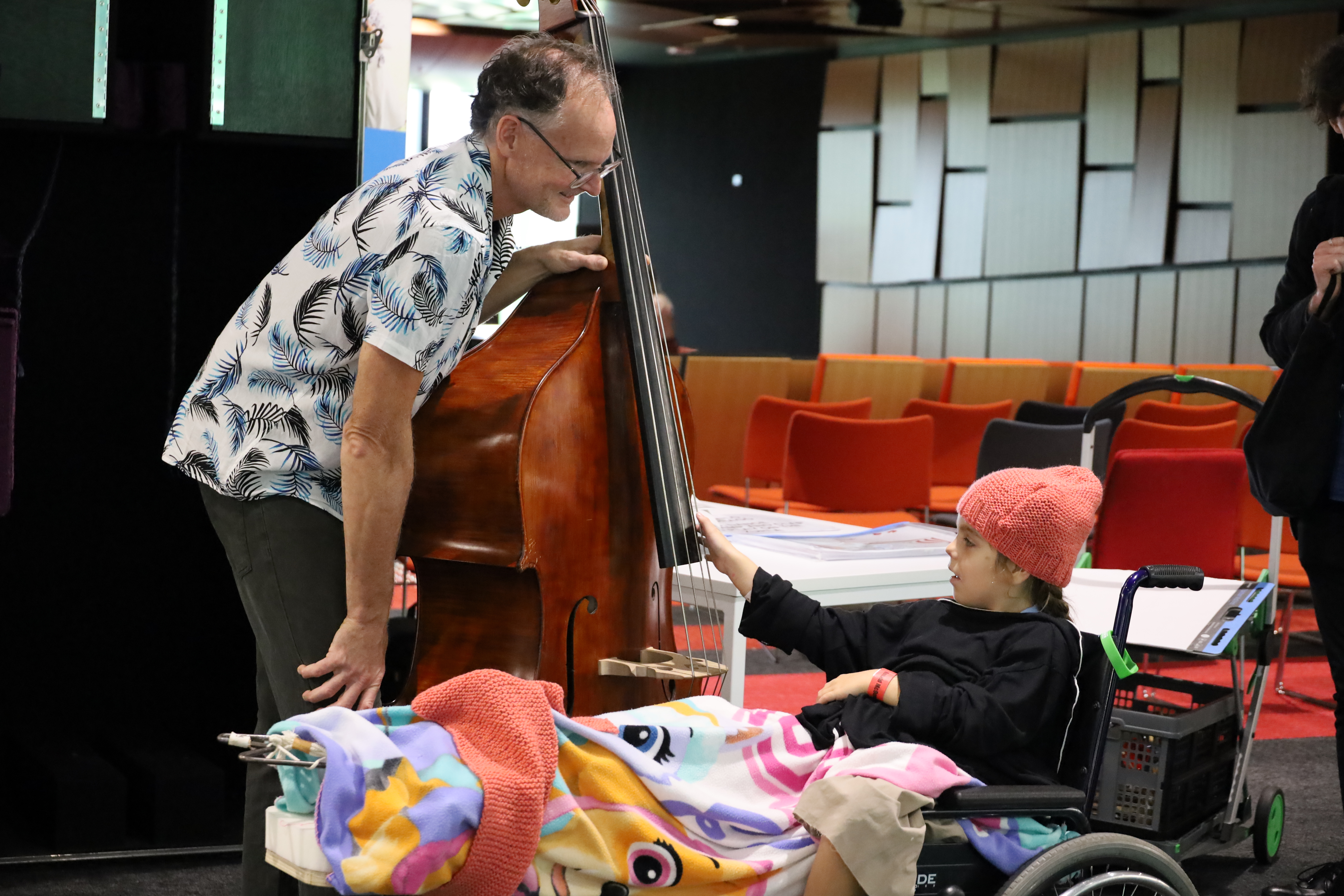 A young girl in a wheel chair reaches out to the strings of a double bass that is being held by a musician