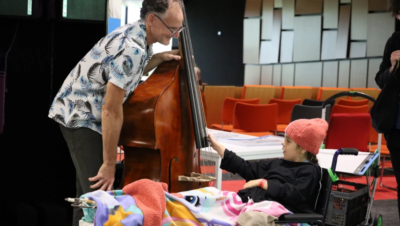 A young girl in a wheel chair reaches out to the strings of a double bass that is being held by a musician
