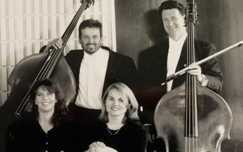 Double bass players pose for a group photo in formal concert attire, in front of doors to a concert hall auditorium.